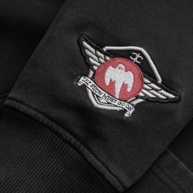 Close-up of a black hoodie sleeve featuring an embroidered winged ghost emblem with a red circle, white bird, and the words 'The show must go on'.