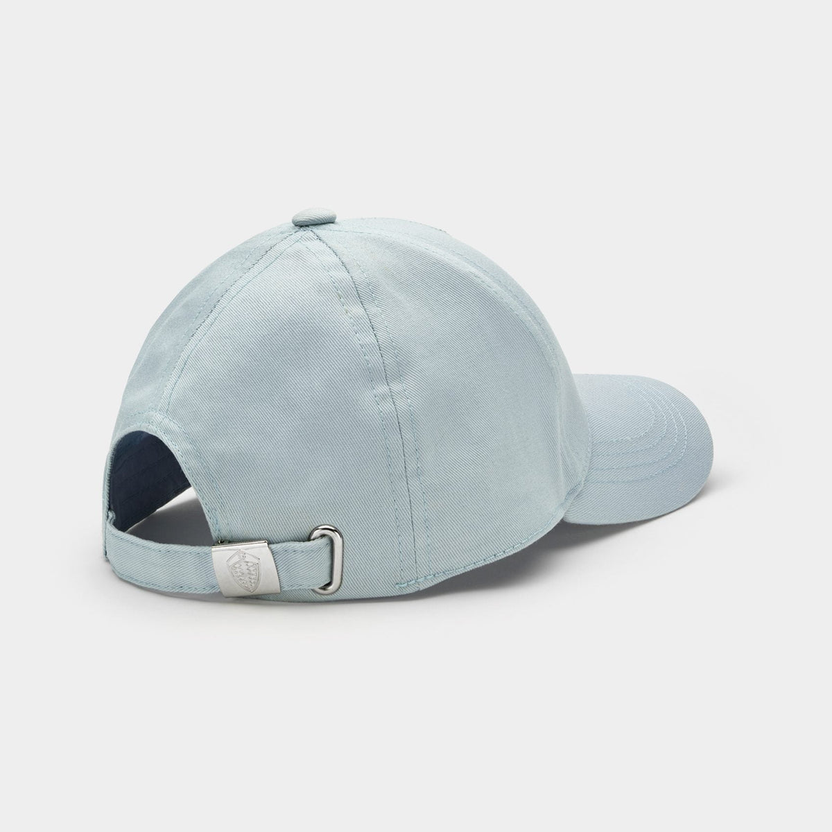A light blue cotton cap shown from a rear three-quarter angle, featuring an adjustable fabric strap with a silver metal buckle, set against a clean white background.