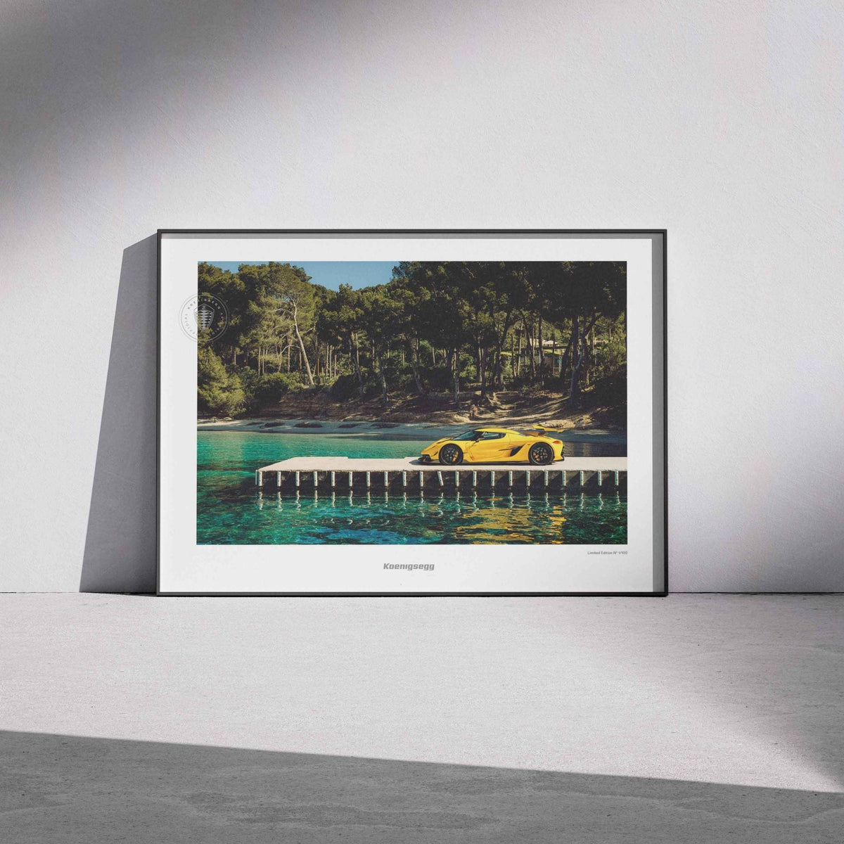 A framed art print leaning against a white wall on a gray floor. The print shows a yellow Jesko on a dock over turquoise water with a forest background.
