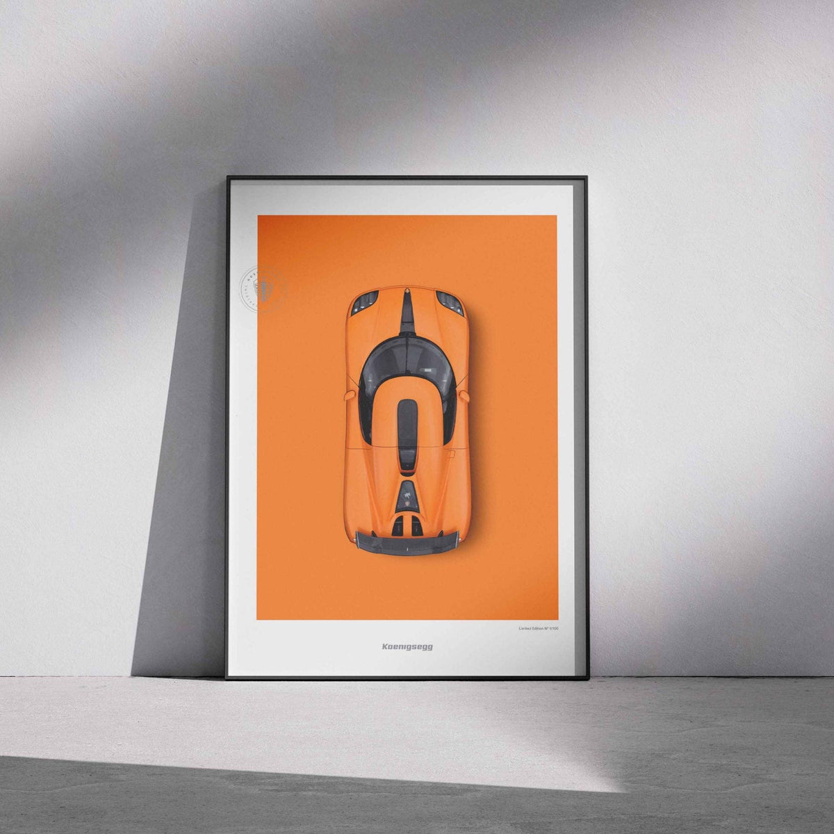 A framed art print featuring a top-down orange CCXR on an orange background, leaning against a light gray wall on a concrete floor with dramatic shadows.