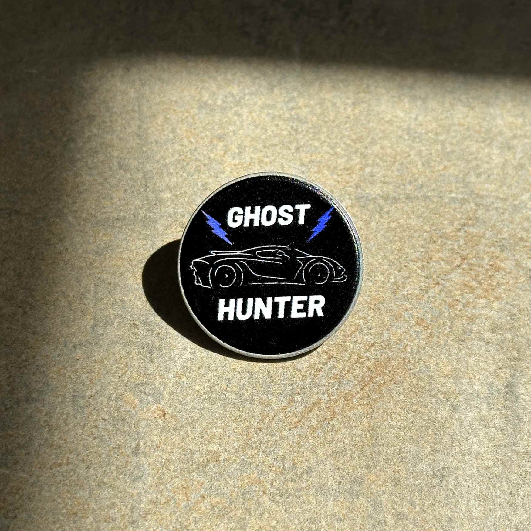 A circular black pin featuring a white outline of a sports car, blue lightning bolts, and the words 'GHOST HUNTER' in white, placed on a textured beige surface with dramatic shadows.