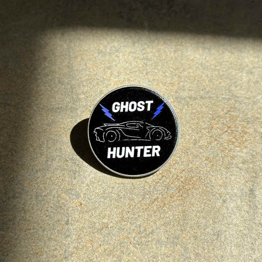 A circular black pin featuring a white outline of a sports car, blue lightning bolts, and the words 'GHOST HUNTER' in white, placed on a textured beige surface with dramatic shadows.