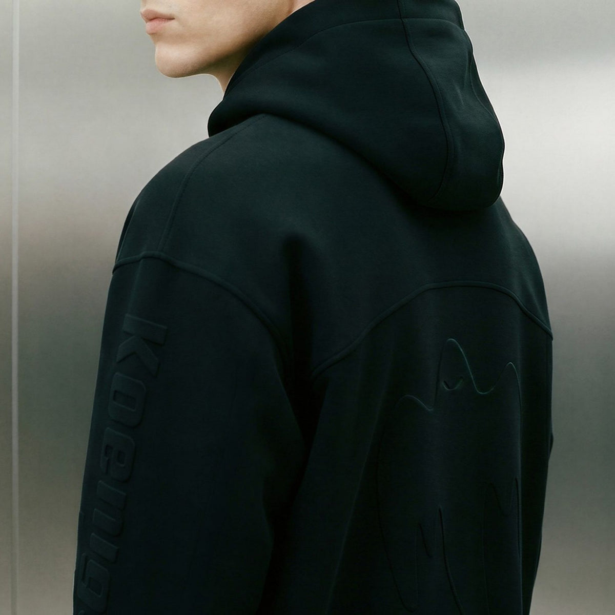 A close-up side view of a person wearing a black Koenigsegg hooded sweatshirt with tonal embroidered details, including vertical text on the sleeve and abstract embroidery on the back, against a gray metallic background.