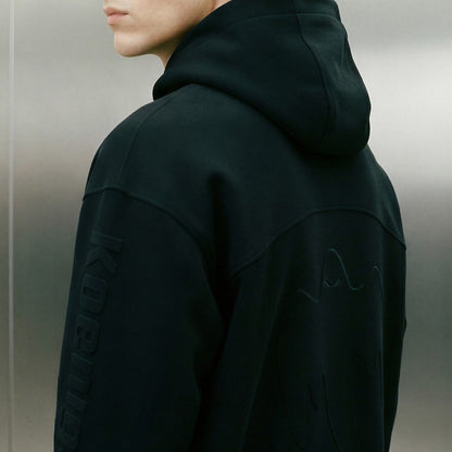 A close-up side view of a person wearing a black Koenigsegg hooded sweatshirt with tonal embroidered details, including vertical text on the sleeve and abstract embroidery on the back, against a gray metallic background.