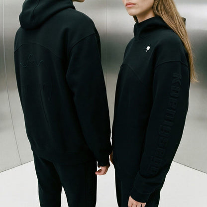 Two models wearing black Koenigsegg hooded sweatshirts. The model on the left shows the back of the hoodie with tonal graphic embroidery, and the model on the right shows the front three-quarter view with 'KOENIGSEGG' printed vertically on the sleeve and a small white ghost logo on the chest.