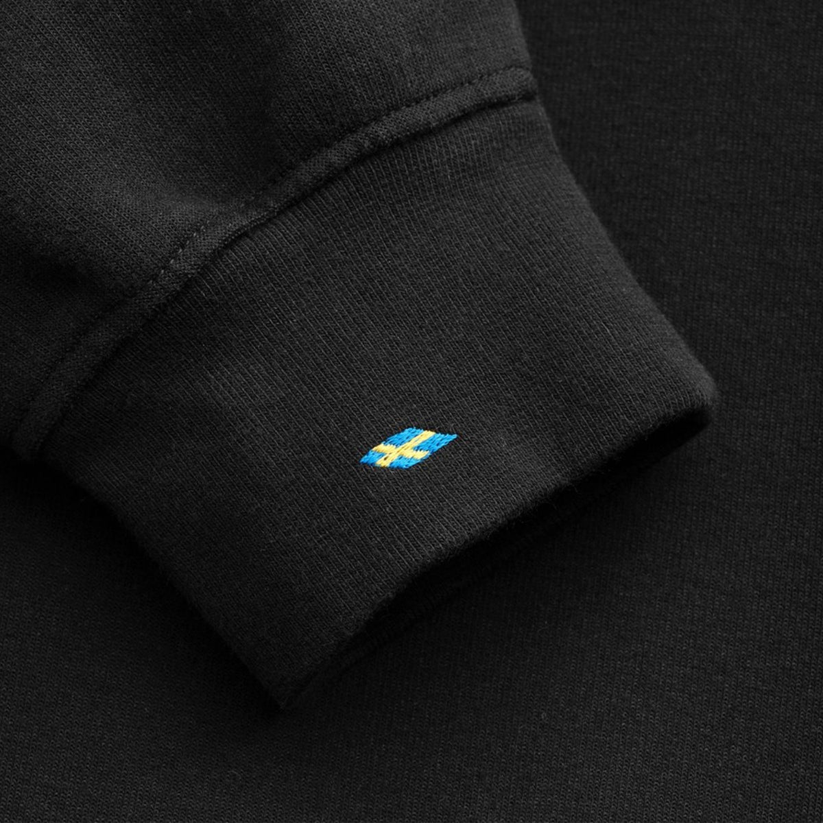 Close-up of a black ribbed knit cuff with a small embroidered blue and yellow Swedish flag logo on the side.