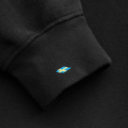 Close-up of a black ribbed knit cuff with a small embroidered blue and yellow Swedish flag logo on the side.