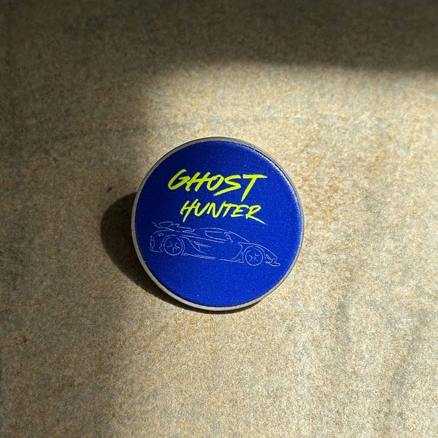 A round enamel pin with a blue background featuring the yellow text 'GHOST HUNTER' and a white outline drawing of a sports car, resting on a textured beige surface.