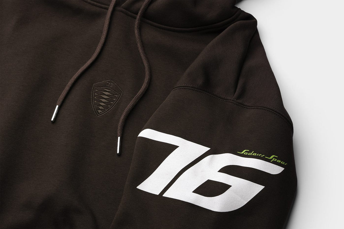 Close-up of a dark brown hoodie with a large white '76' graphic on the sleeve, a small green script logo, and a tonal shield patch on the chest.