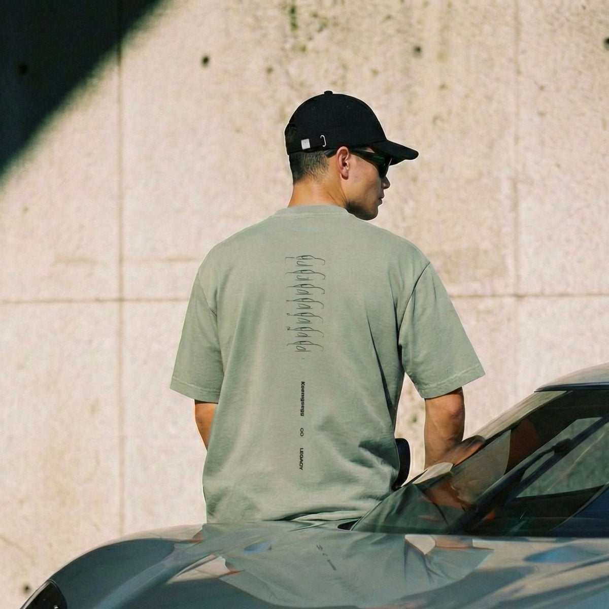 A man wearing a light sage green t-shirt with a vertical graphic print on the back, a black baseball cap, and sunglasses, standing next to a car in front of a textured concrete wall.