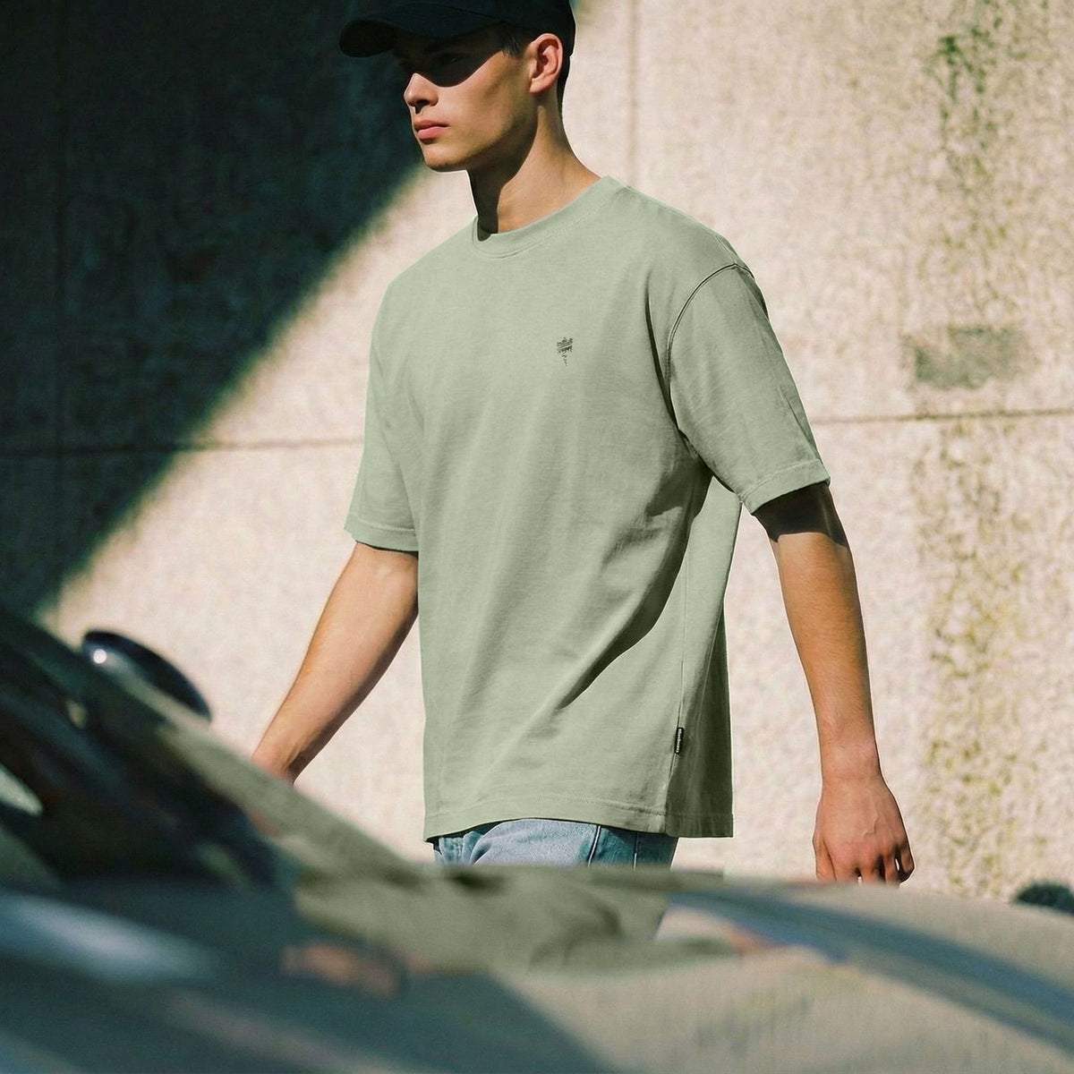 A man wearing a light sage green crewneck t-shirt and a black baseball cap, standing outdoors in front of a textured wall with strong sunlight and shadows.