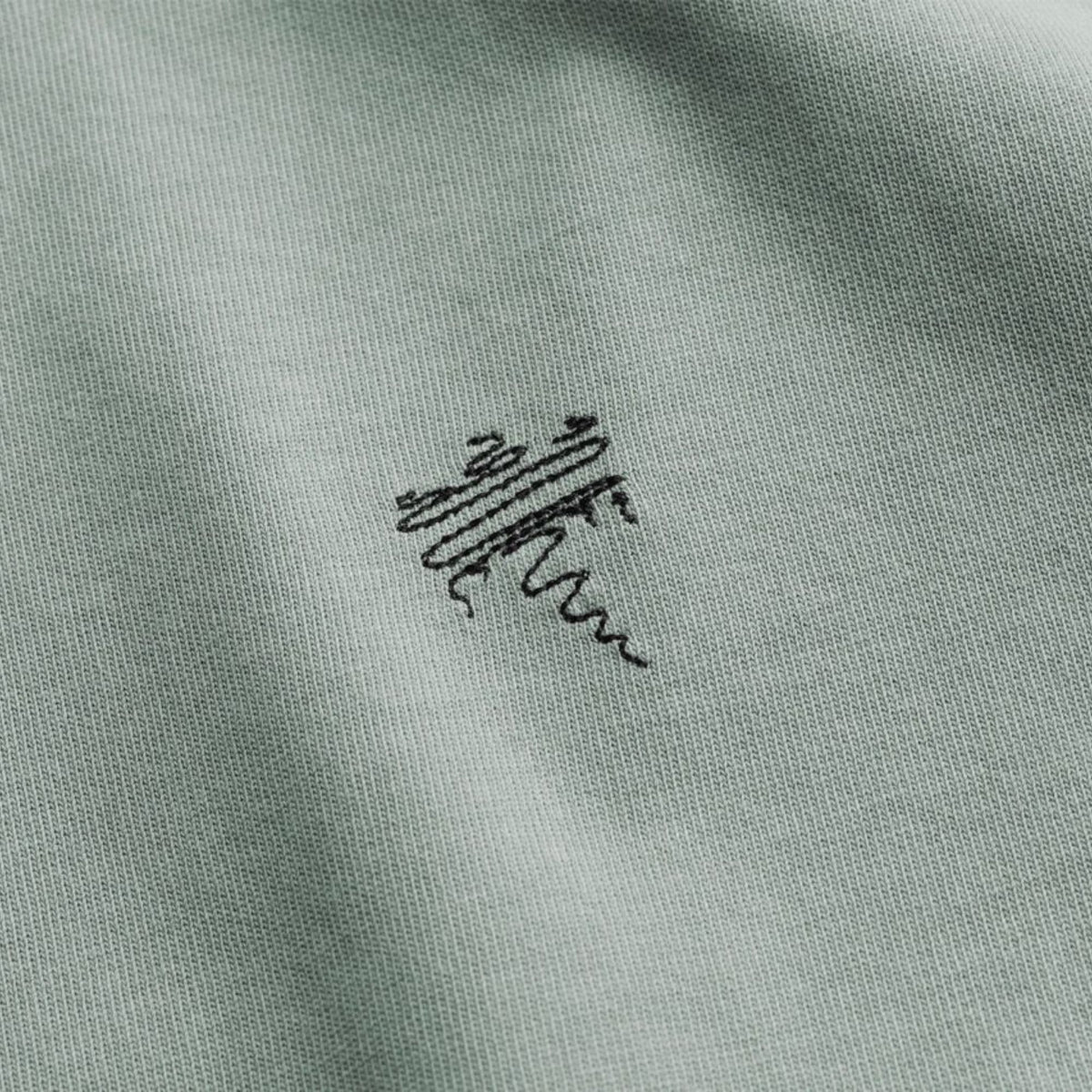 Close-up of a light sage green knit fabric with a small black embroidered Koenigsegg logo in the center.