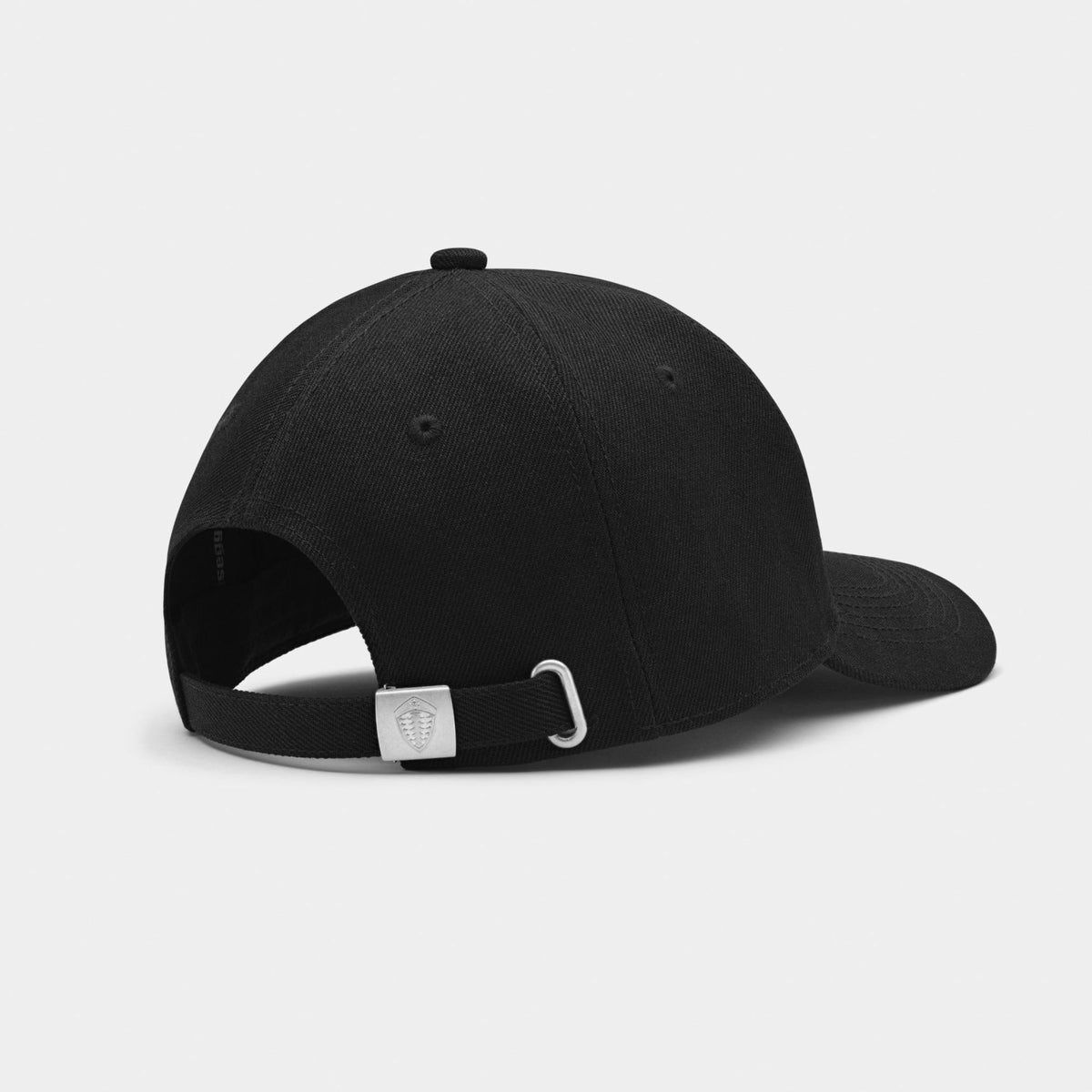 Black baseball cap shown from the back, featuring a curved brim, black adjustable strap, and a silver metal buckle with a small shield logo on a light gray background.