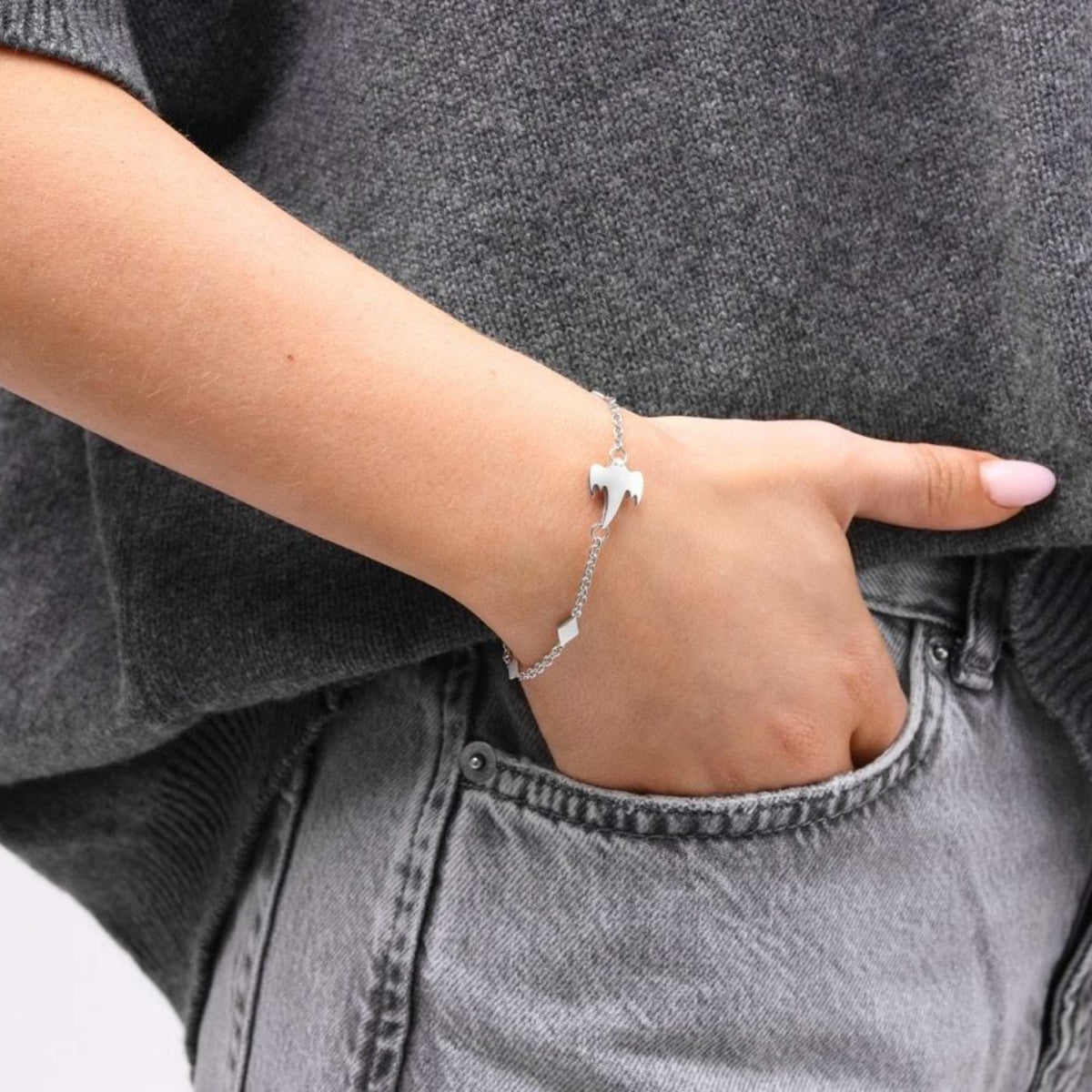 A close-up of a woman's hand resting in a gray denim pocket, wearing a delicate silver Koenigsegg chain bracelet with a small silver ghost charm.