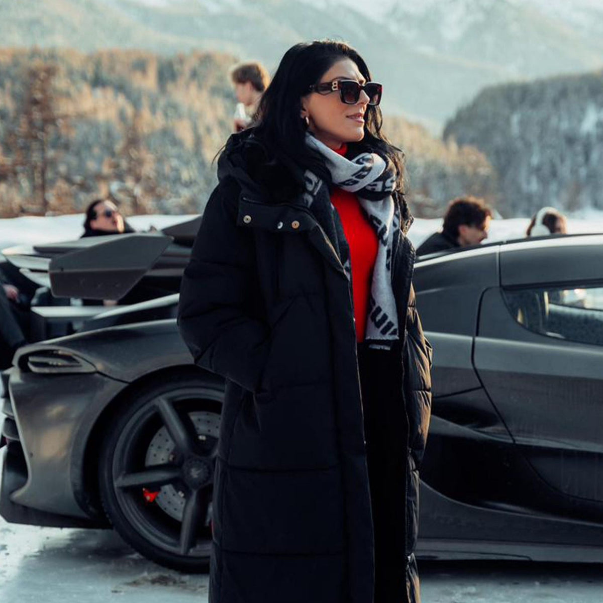 A woman wearing a long black puffer coat, red sweater, gray Koenigsegg Gear patterned scarf, and sunglasses stands in front of a Koenigsegg in a snowy outdoor setting.