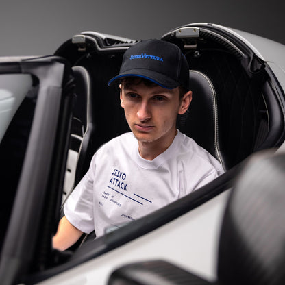 A man sitting inside a white and black Koenigsegg car, wearing a black baseball cap with blue embroidery reading 'SuperVettura' and a white t-shirt with blue text reading 'JESKO ATTACK'.