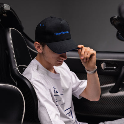 A man sitting in a Koenigsegg car interior wearing a black baseball cap with blue 'SuperVettura' embroidery and the Technical Kinetic SuperVettura Tshirt.