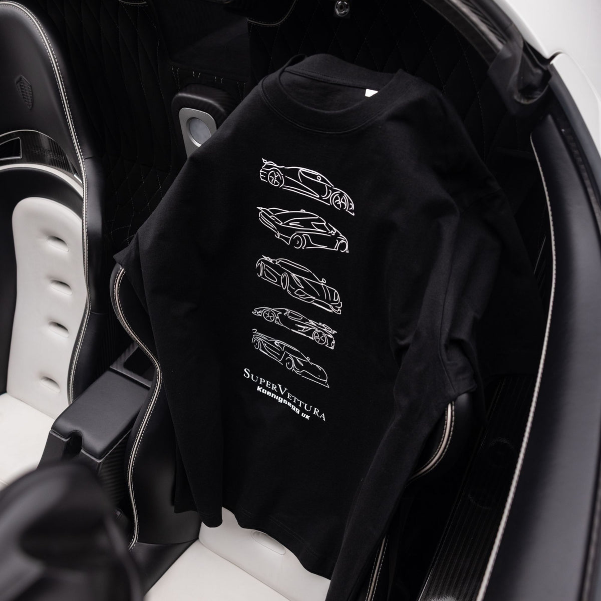A black crewneck sweatshirt featuring white line-art illustrations of sports cars and the text 'SUPERVETTURA' printed on the back, draped over the seat of a Koenigsegg car interior.