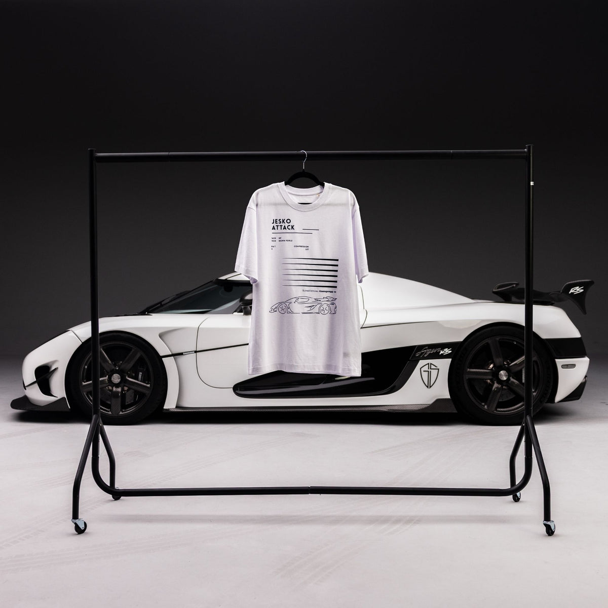 A white graphic SuperVettura t-shirt with black text and a car illustration hanging on a black metal clothing rack, positioned in front of a white and black Koenigsegg car against a dark studio background.