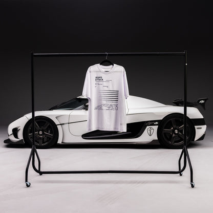 A white graphic SuperVettura t-shirt with black text and a car illustration hanging on a black metal clothing rack, positioned in front of a white and black Koenigsegg car against a dark studio background.
