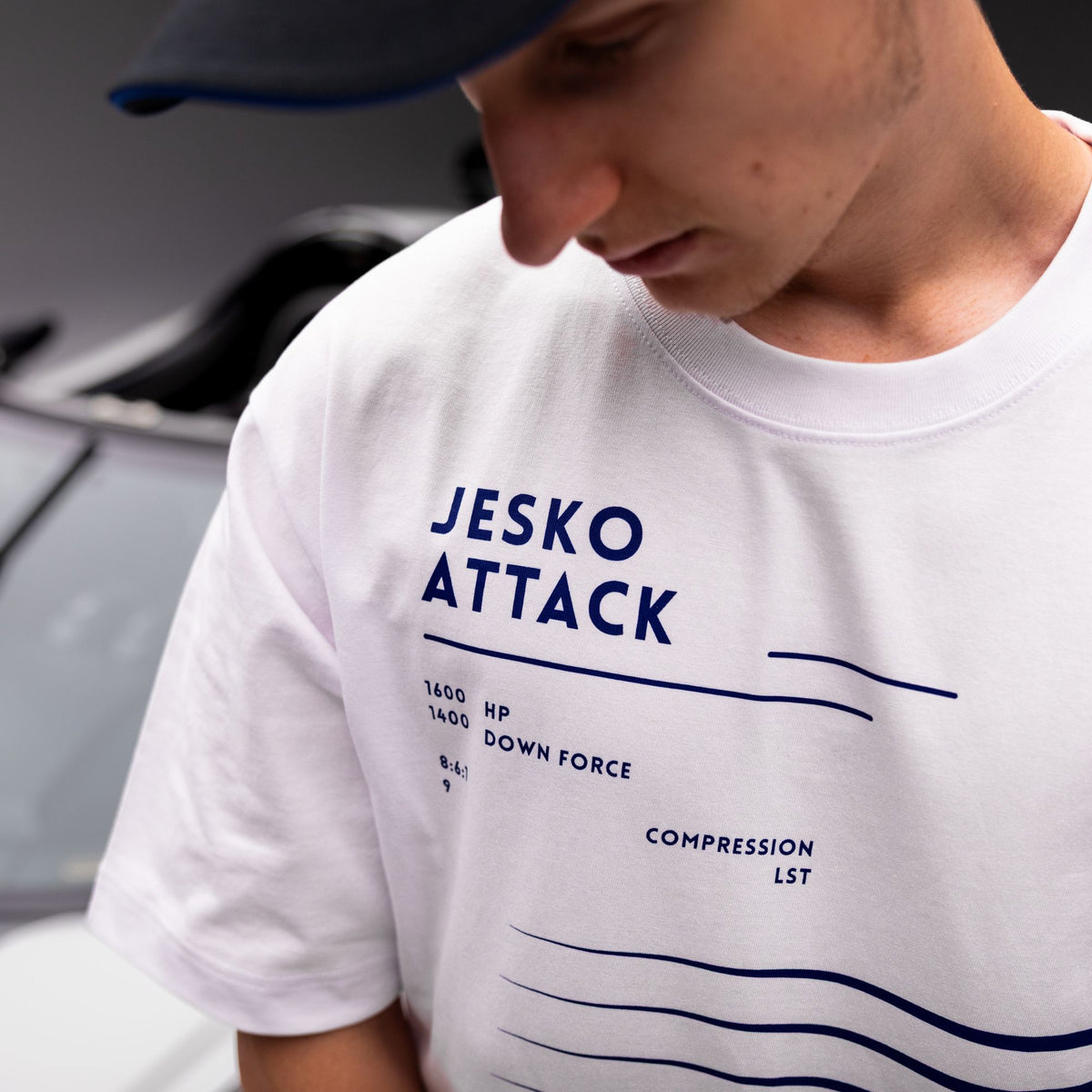 A close-up of a person wearing a white crewneck t-shirt with dark blue printed text and graphics reading 'JESKO ATTACK' and technical specifications, along with a dark blue baseball cap.