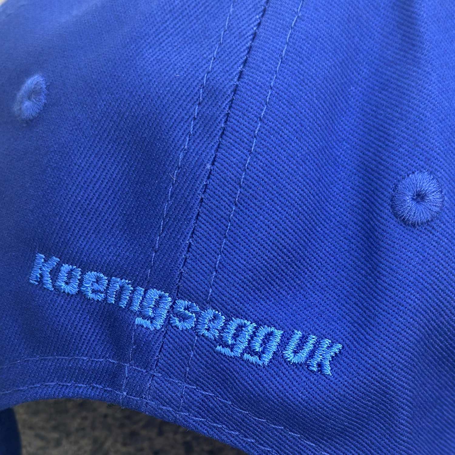 Close-up of a royal blue fabric cap with light blue embroidered text reading 'Koemigsegguk' and matching blue eyelets.