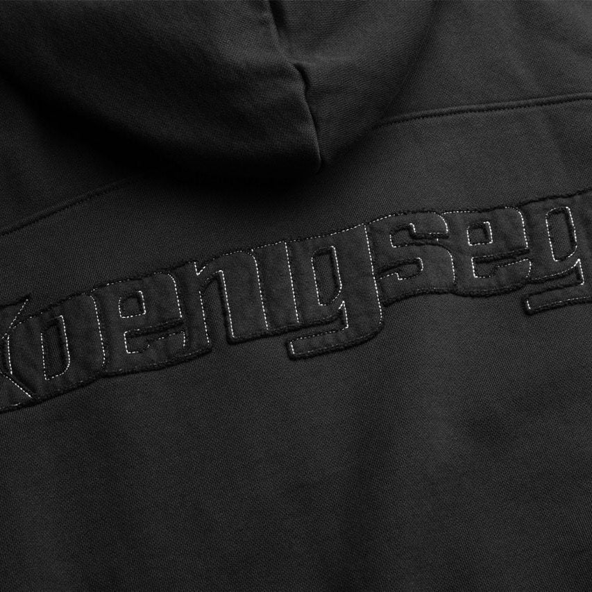 Close-up of a black hoodie back featuring large tonal black embroidered text reading 'Koenigsegg' across the upper back.