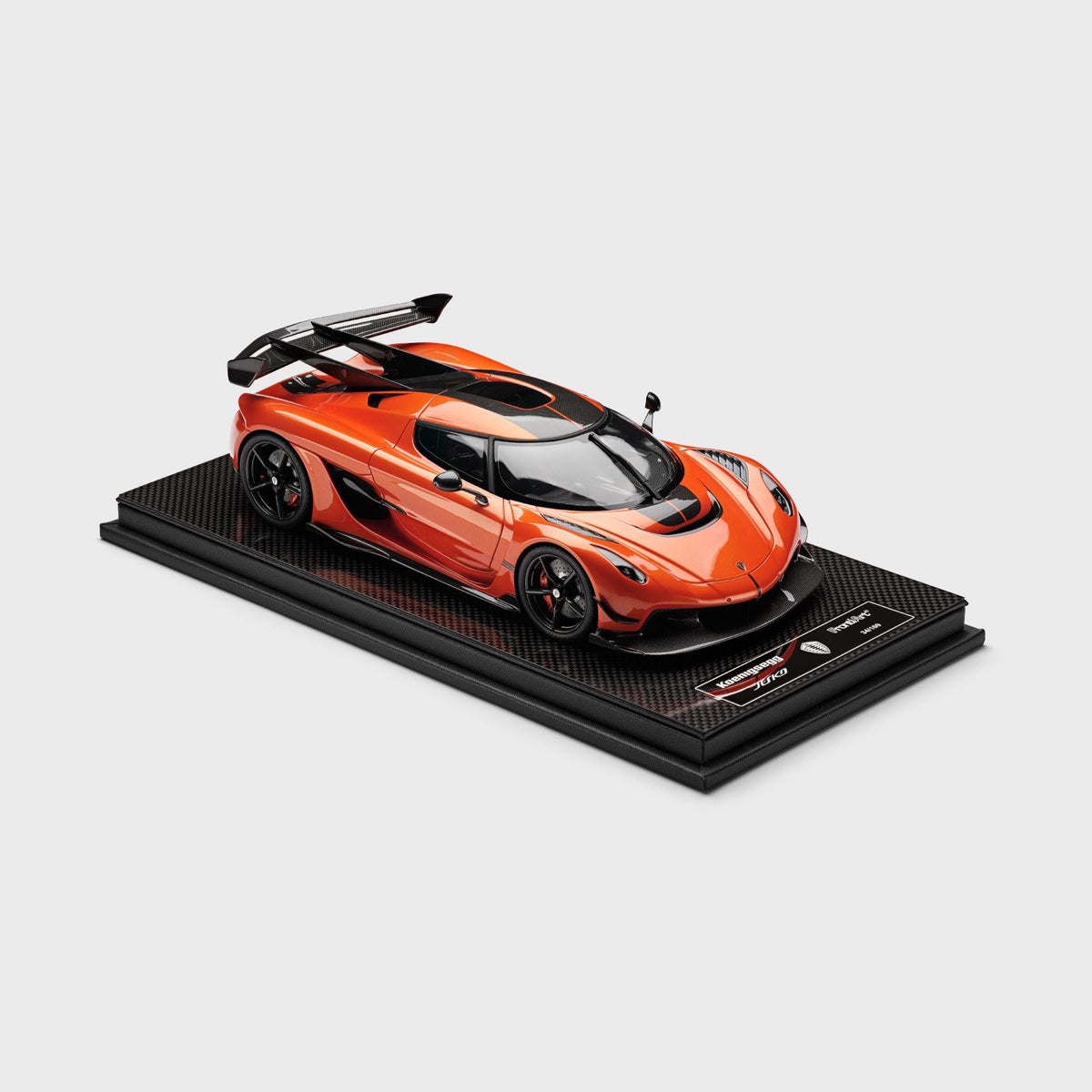 An orange and black scale model of a Jesko Attack, displayed on a black carbon-fiber-textured base with a small plaque, set against a solid light gray background.