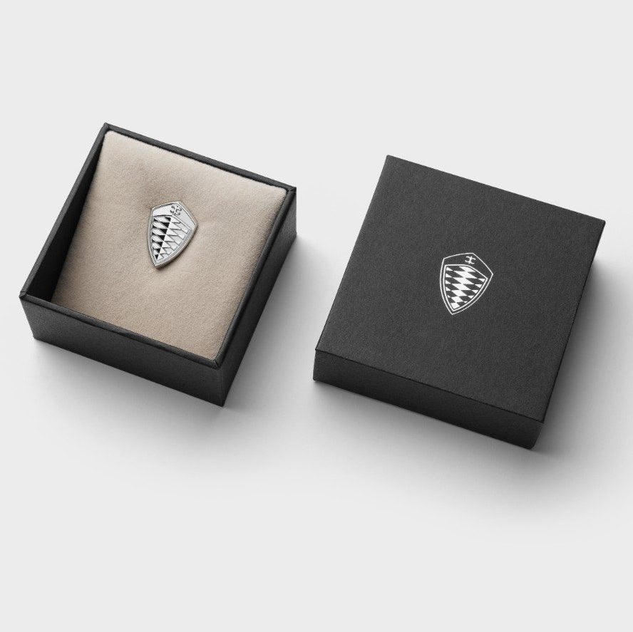 A silver shield-shaped pin badge displayed in an open black presentation box with a beige insert, alongside a closed black box featuring the same shield logo, on a light gray background.